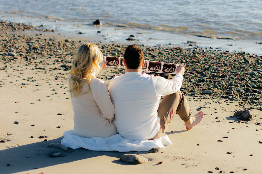 Couple on beach viewing ultrasound images, celebrating pregnancy together for their nh beach maternity session