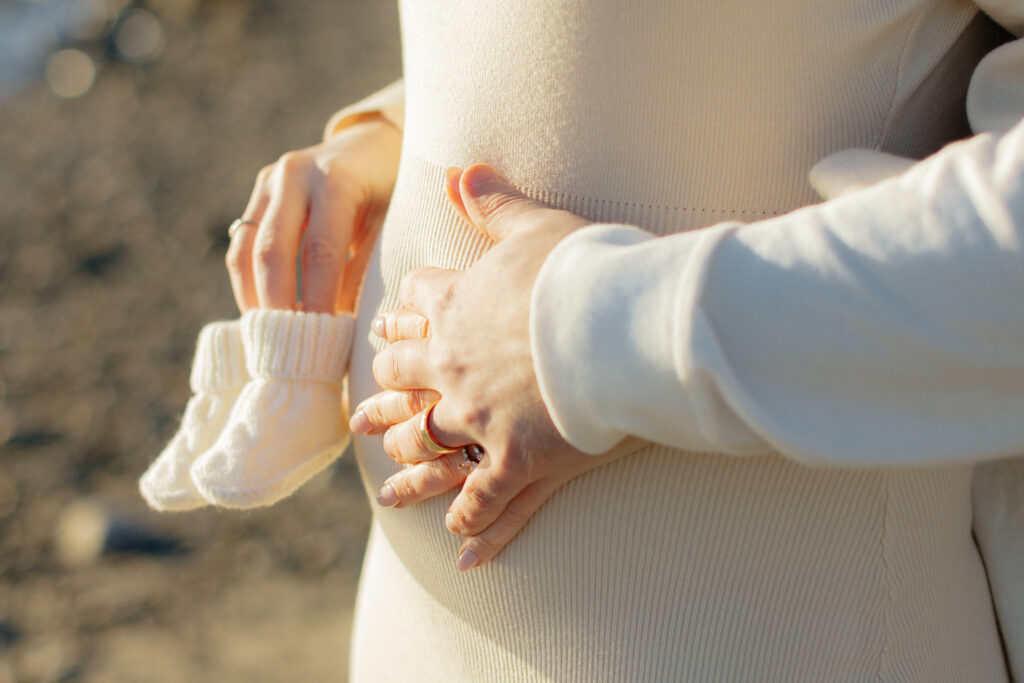 Close-up of hands embracing a pregnant belly, holding baby booties at an nh beach maternity session, symbolizing a loving expectation for a new arrival.