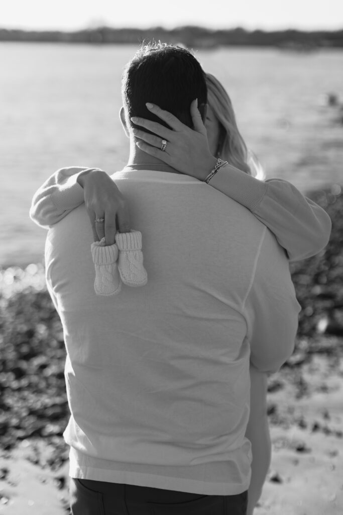 Couple embracing by water, holding baby booties, symbolizing pregnancy announcement.