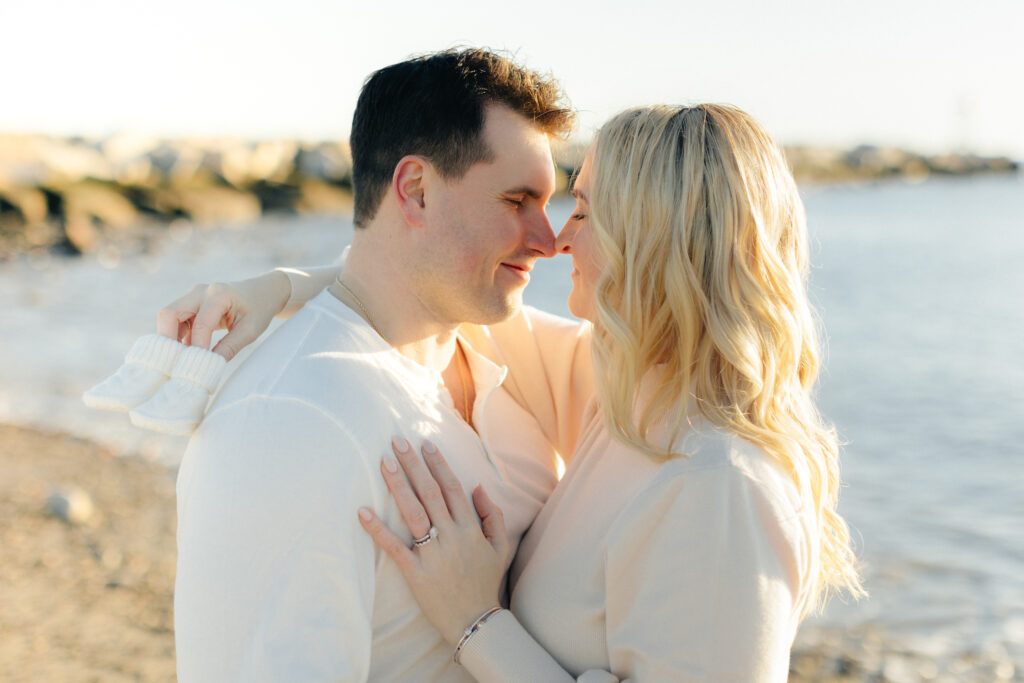 Couple embracing by the beach, holding baby shoes, symbolizing love and anticipation for their new arrival.