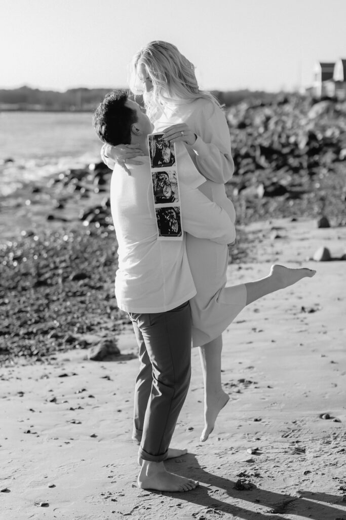 Couple celebrating pregnancy by the beach with ultrasound photos, embracing joy and love under the sun.