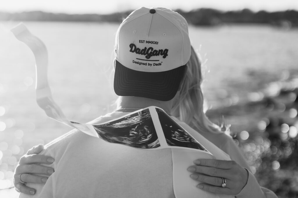 Man in DadGang cap holds ultrasound by the water, embraced by woman with ring.