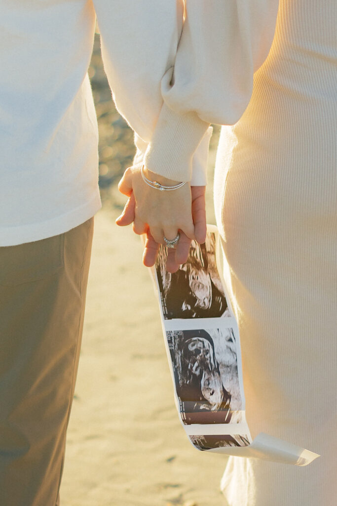 Couple holding hands with pregnancy ultrasound images; celebration of new beginnings on a sunny day at an nh beach maternity session.