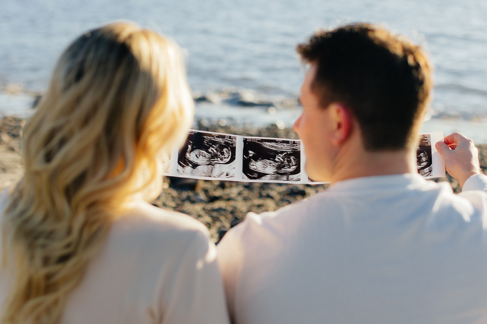 Couple by the beach holding ultrasound images, celebrating pregnancy announcement, with ocean in the background.