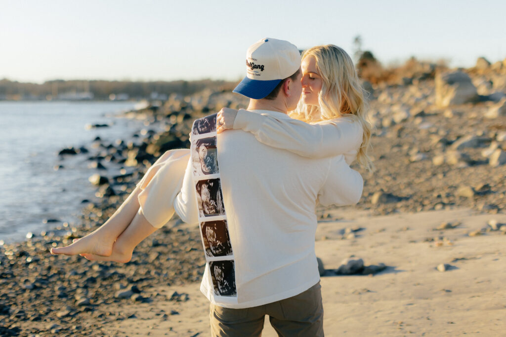 Couple embraces on rocky beach, holding ultrasound photos, celebrating pregnancy under sunny sky at their beach maternity session.