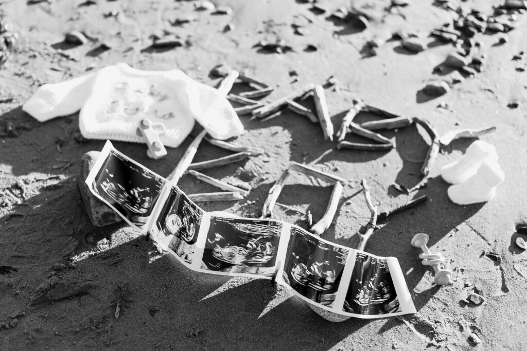 Ultrasound photos on sand with baby clothes and sticks spelling BABY at a creative nh beach maternity session.