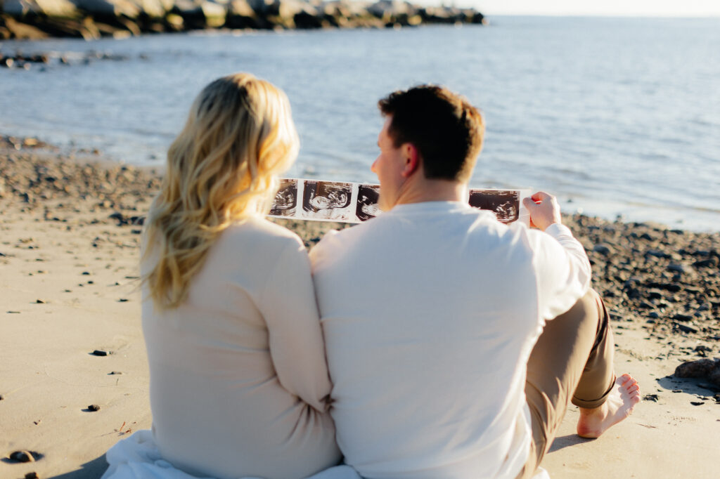 Couple sitting on beach holding ultrasound photos, joyful moment by the sea at their nh beach maternity session.