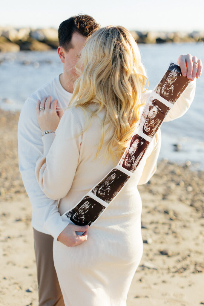 Couple embracing on beach holding ultrasound photos, celebrating pregnancy.