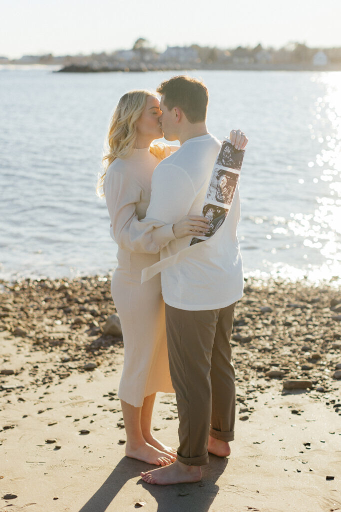 Couple kissing on beach holding baby ultrasound strip, captured in soft sunlight in their nh beach maternity session.
