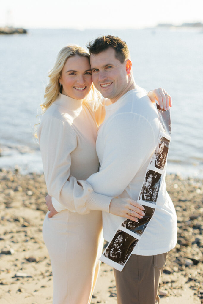 Couple smiling by the beach holding ultrasound images, celebrating pregnancy announcement through a nh beach maternity session.