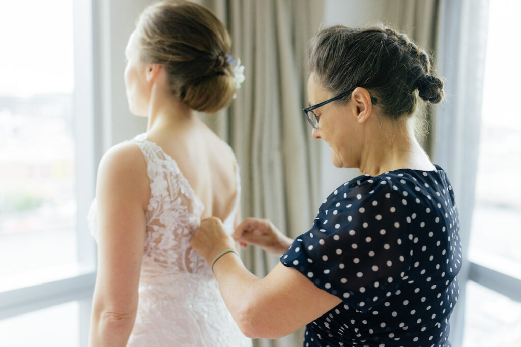 Woman helps bride with lace wedding dress near window.