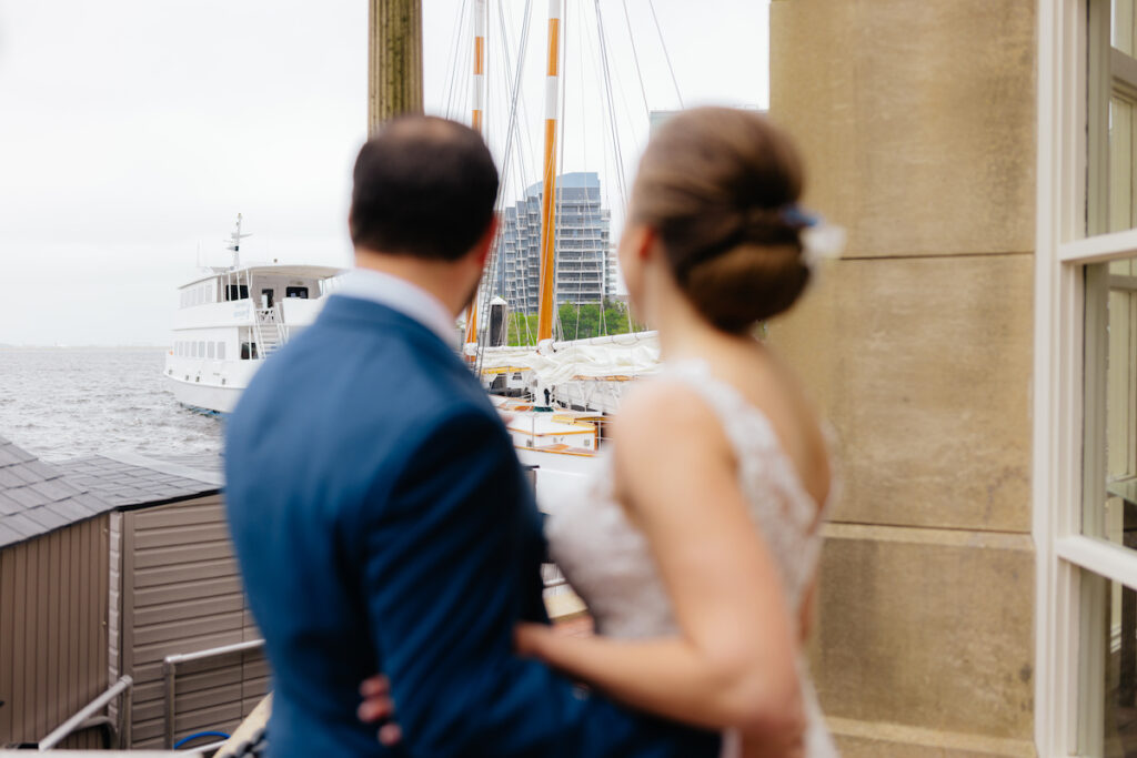 Couple in wedding attire looking at a boat on a waterfront, with city buildings in the background.