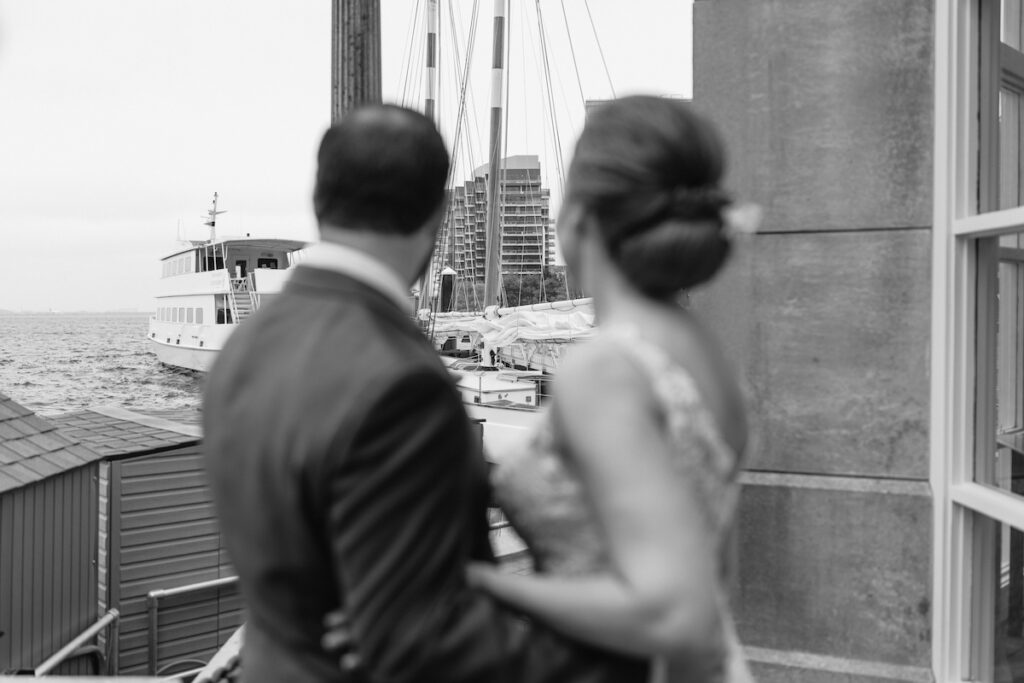 Couple gazes at docked ship in the harbor, with city buildings and ocean view in the background.