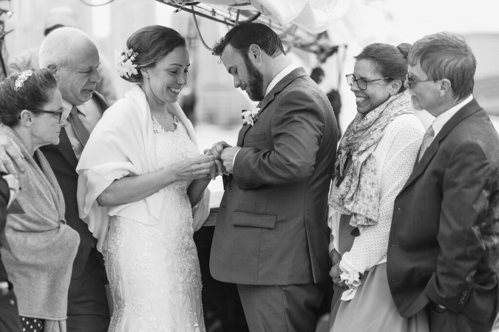 Bride and groom exchanging rings surrounded by family at an outdoor wedding ceremony, smiling and joyful.