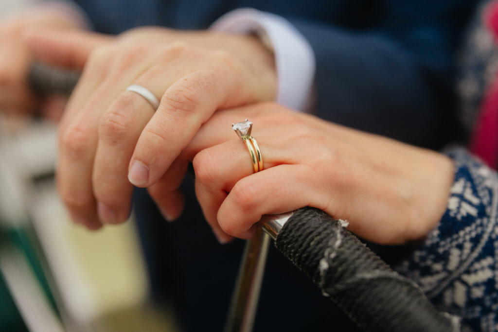 Hands holding each other with engagement and wedding rings on a steering wheel, symbolizing love and commitment.