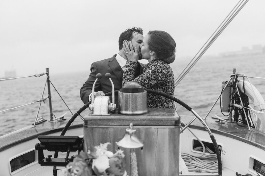 A couple kisses on a classic harbor line sailboat, surrounded by the ocean, under a cloudy sky.