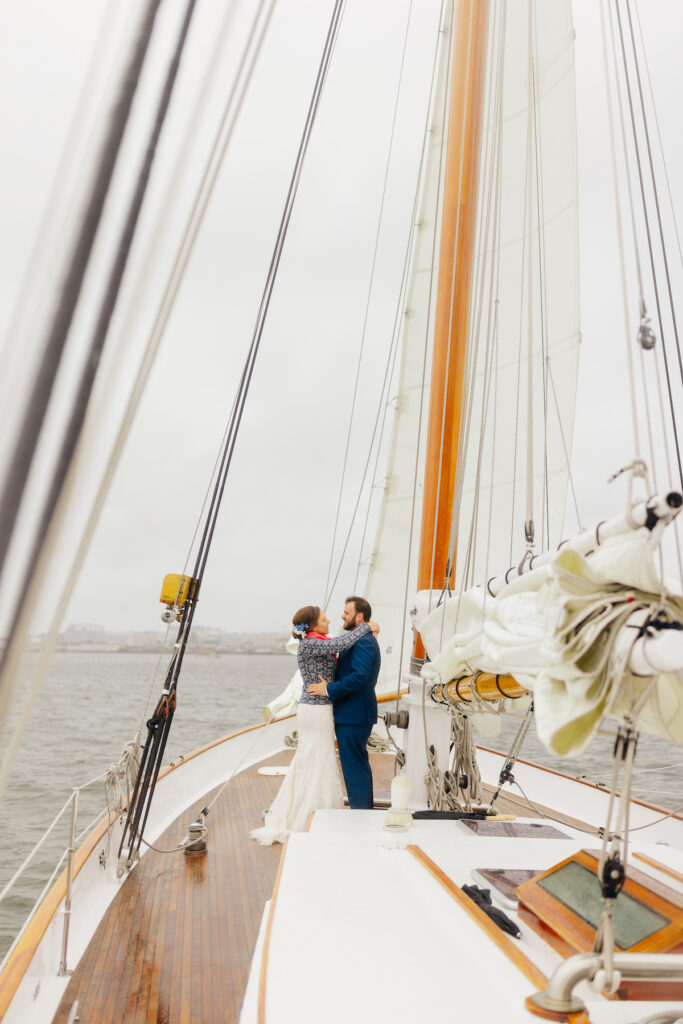 Couple embracing on a classic harbor line sailboat deck, wedding attire, overcast sky, ocean backdrop.