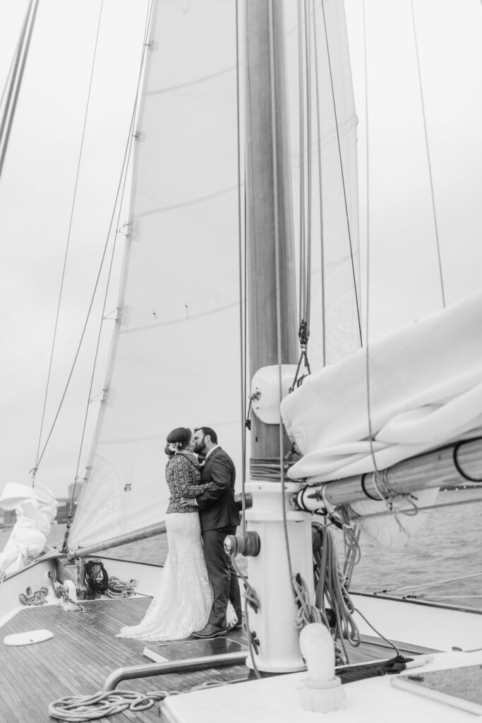 Bride and groom share a romantic kiss on a classic harbor line sailboat under a cloudy sky.