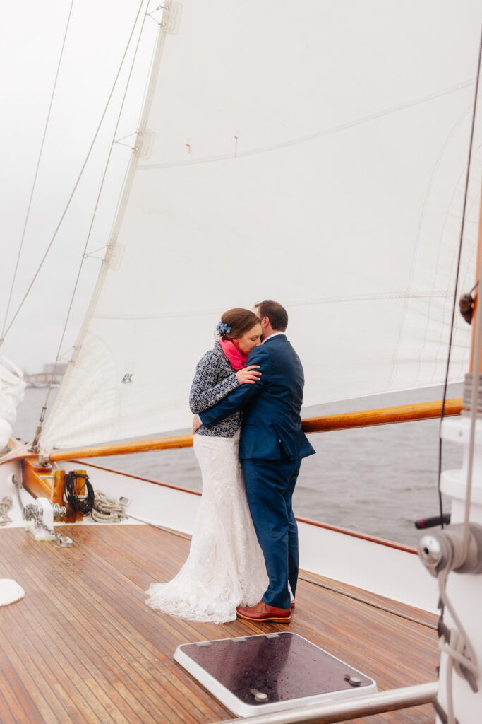 Couple embraces on sailboat deck, bride in lace gown, groom in blue suit, under large white sail on classic harbor line Boston.