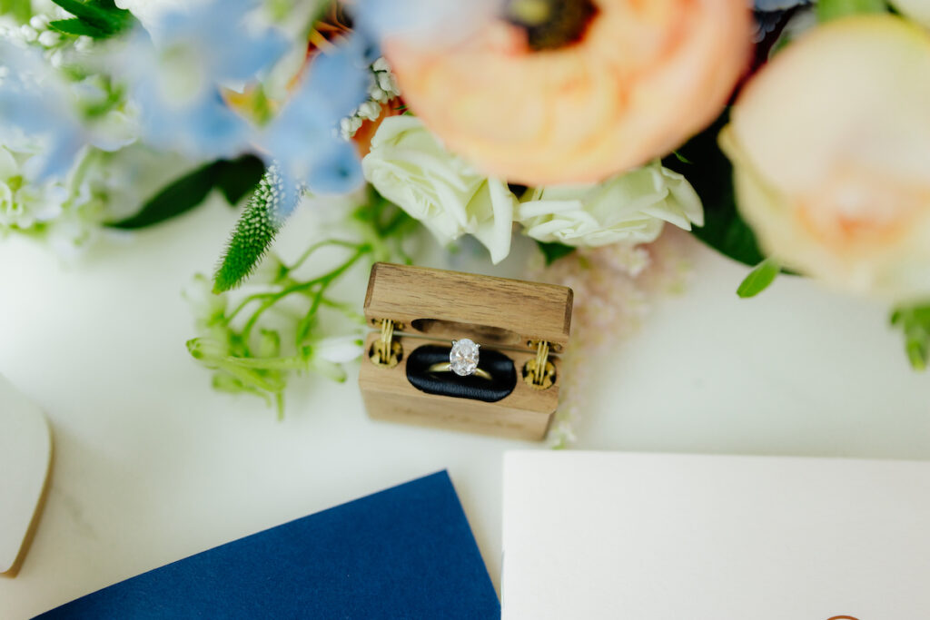 Elegant diamond ring in wooden box with floral bouquet, ideal for weddings or engagements.