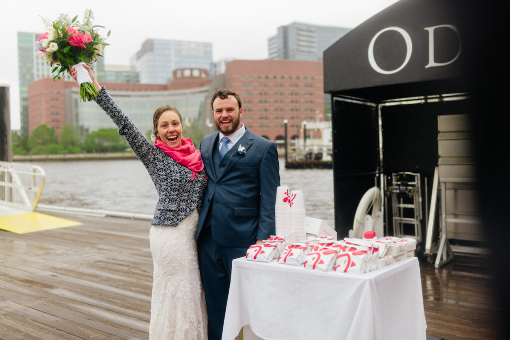 Happy couple celebrating outdoors with bouquet, near river and cityscape, table with snacks displayed in foreground.