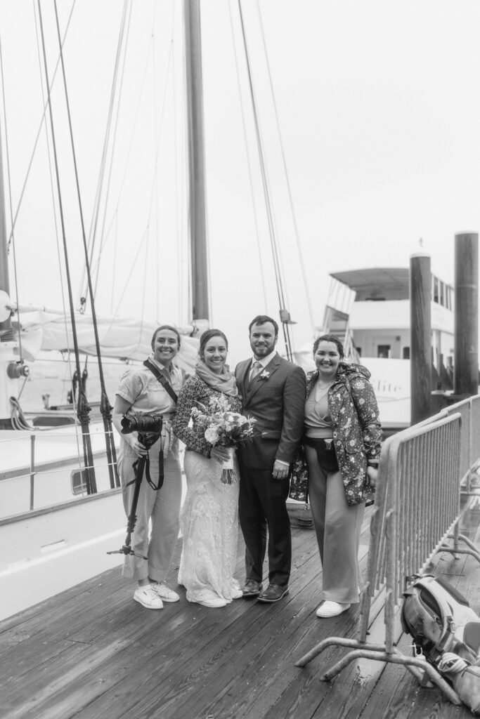 Bride and groom smiling with friends on a docked sailboat, holding flowers, monochrome photo.