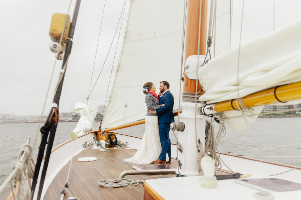 Bride and groom embrace on sailboat deck under cloudy sky, nautical wedding theme.