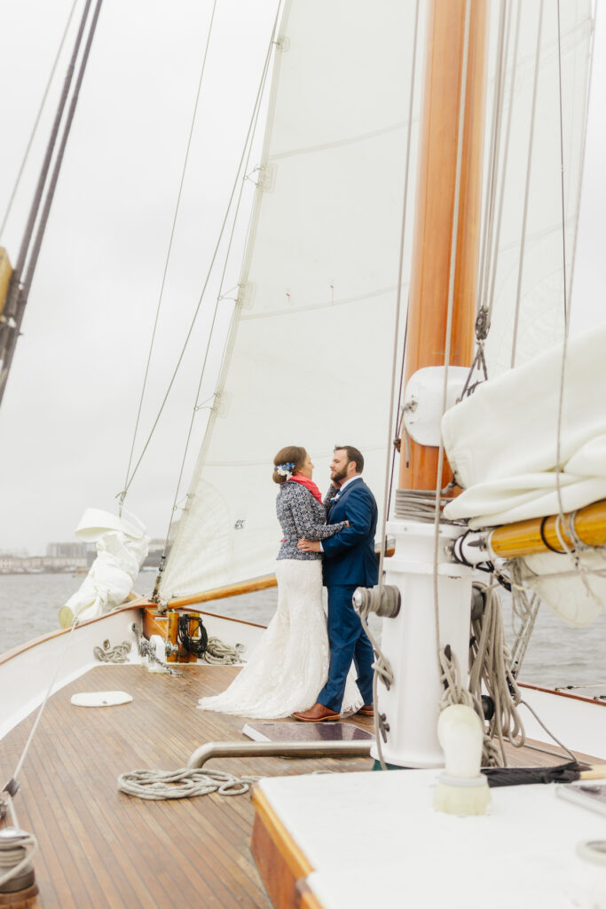 Couple embracing on sailboat, elegant attire, wooden deck, white sails, cloudy sky in background.