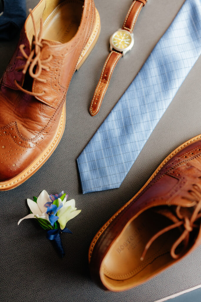 Brown leather shoes, blue tie, leather watch, and boutonniere on gray surface. Perfect for stylish men's fashion.