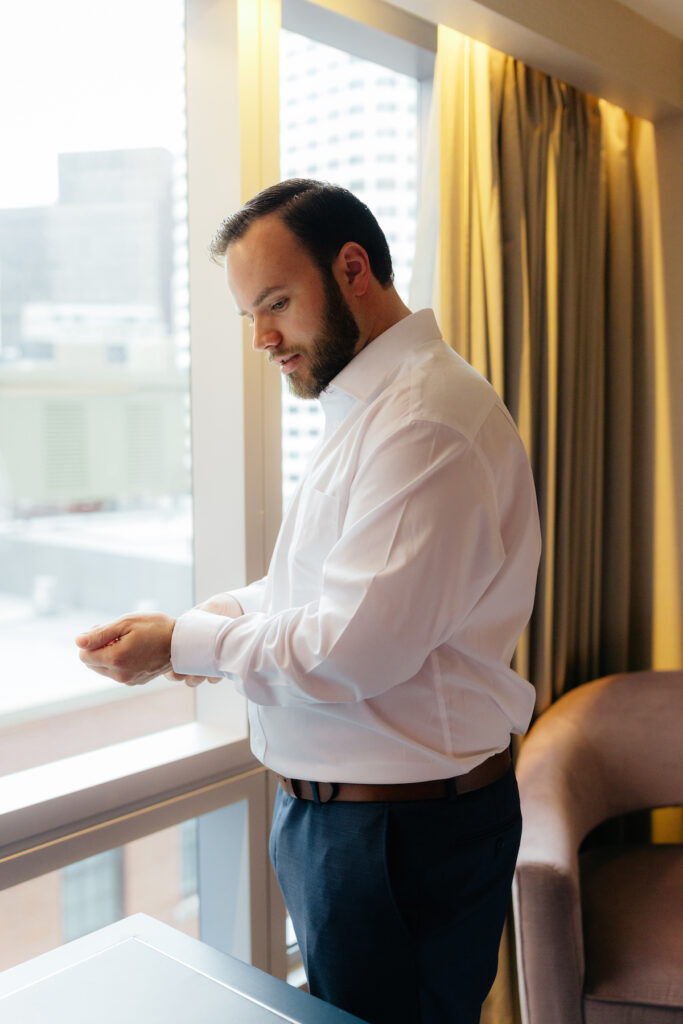 Man in white shirt adjusting cufflinks by a window in a bright room, cityscape in background.